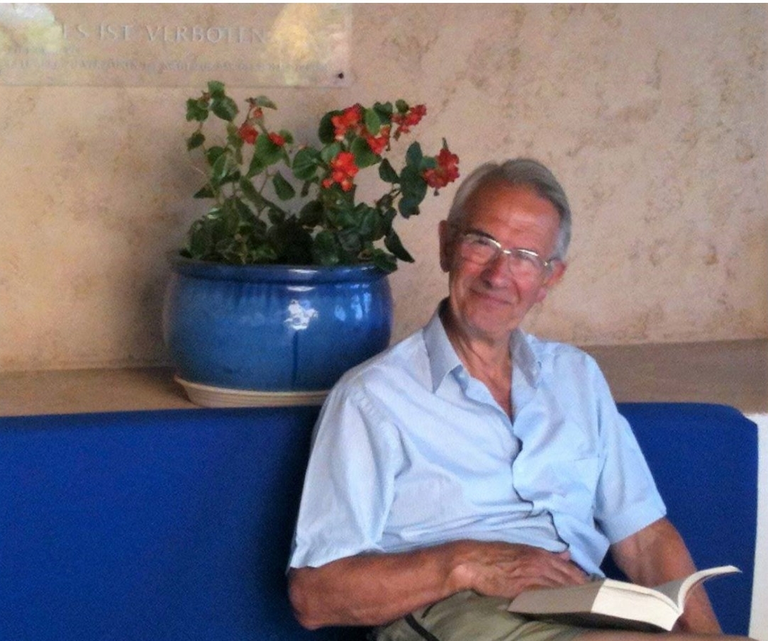 Photograph of composer Christopher Field sitting down, reading a book. On the shelf behind is a blue flower pot with red flowers
