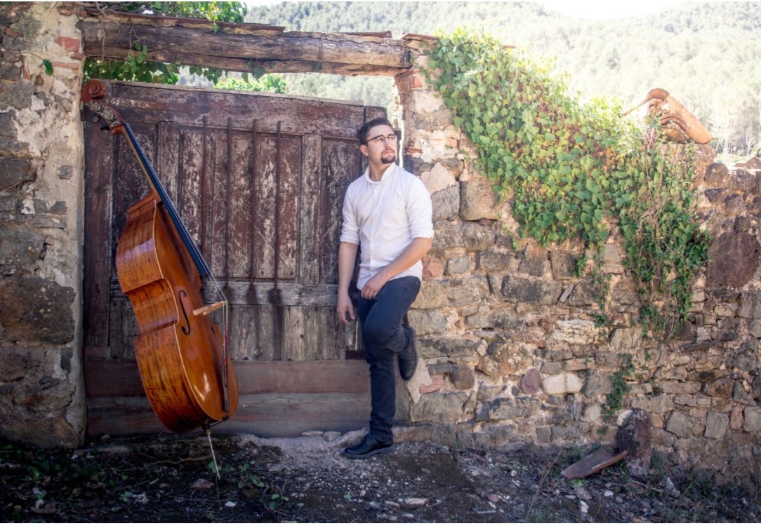 Image of composer Daniel Chiva sanz with is double bass. He is standing againstan old wooden gate in a walled garden .
