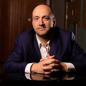 Colour photograph of composer Francis Faux wearing a white shirt and blue jacket with his hands crossed.