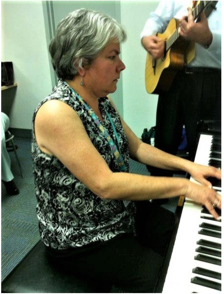 Photographic image of composer Lourdes C Montgomery playing the piano with a guitarist in the background
