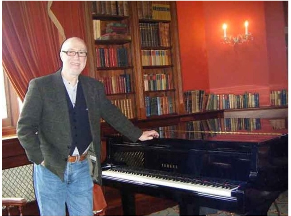 Image of the composer Peter Byrom Smith standing with his hand on a piano