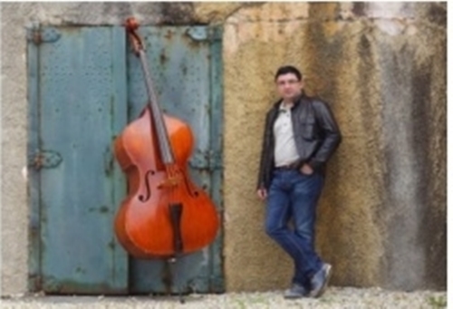 Composer Simon Garcia in a black leather jacket and jeans stands leaning against a wall next to a large, reddish-brown double bass leaning against a teal-colored door.