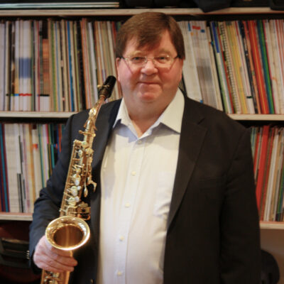 Ohotograph of composer and musician James Rae holding a saxophone with 2 shelves of sheet music behind him,
