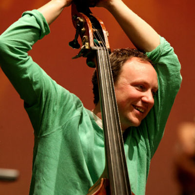 Photo of composer Oli Hayhurst wearing a green shirt and holding the neck of a double bass.