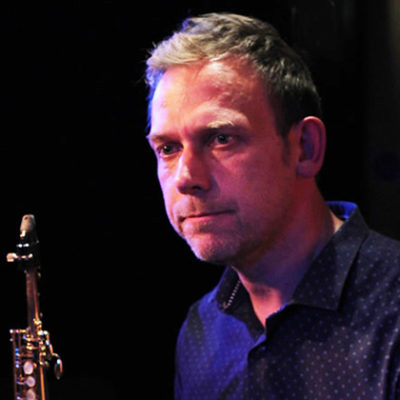 Photo of composer Rob Hall wearing a dark blue shirt and tie, holding a clarinet.