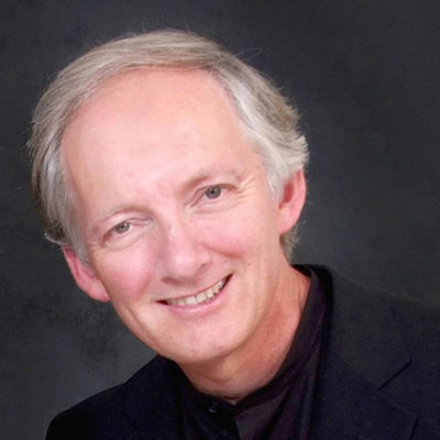 Photograph of composer Robin Ireland who has grey hair and is wearing a black shirt and dark jacket.