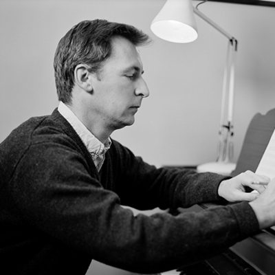 Black and white image of composer Philip Godfrey, . He is sat at a piano and appears to be working on a manuscript.